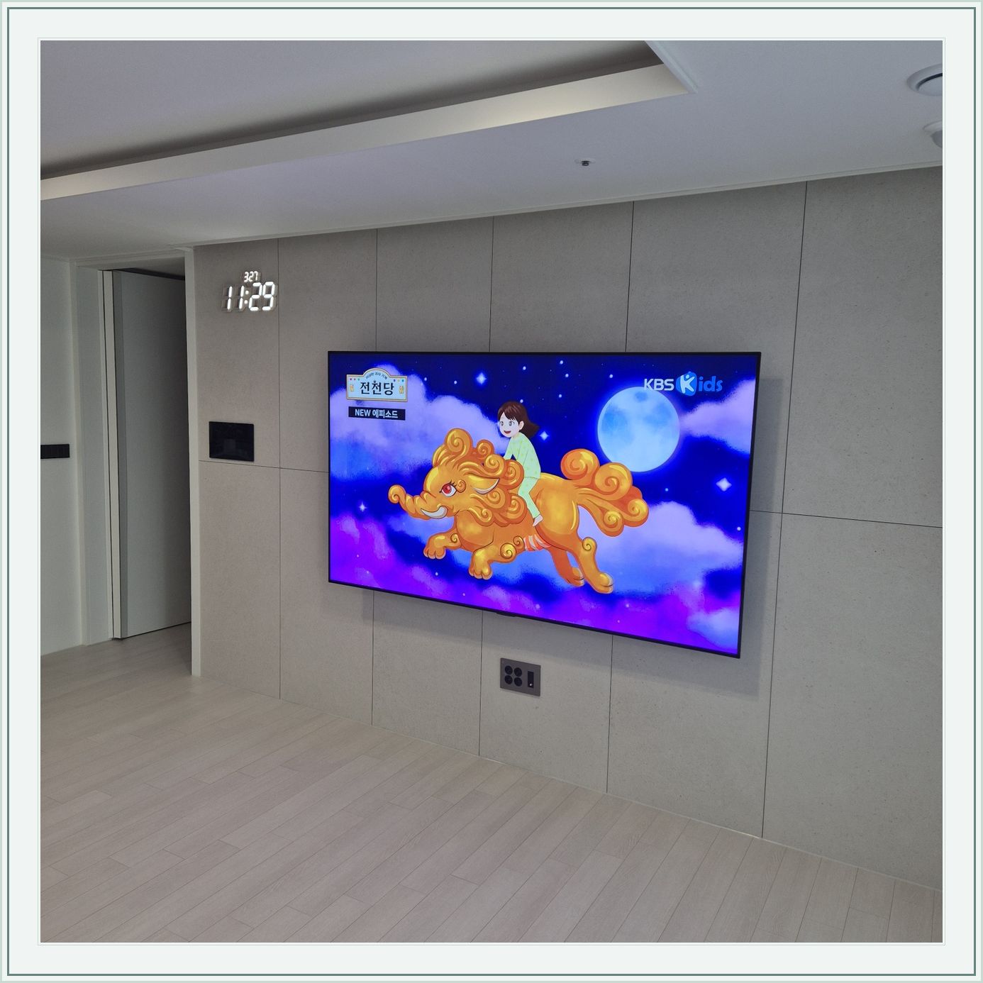 회색 판넬 벽 위의 화면과 디지털 시계, 벽면 패널 이음선과 TV 중심의 배치를 비교할 수 있습니다
