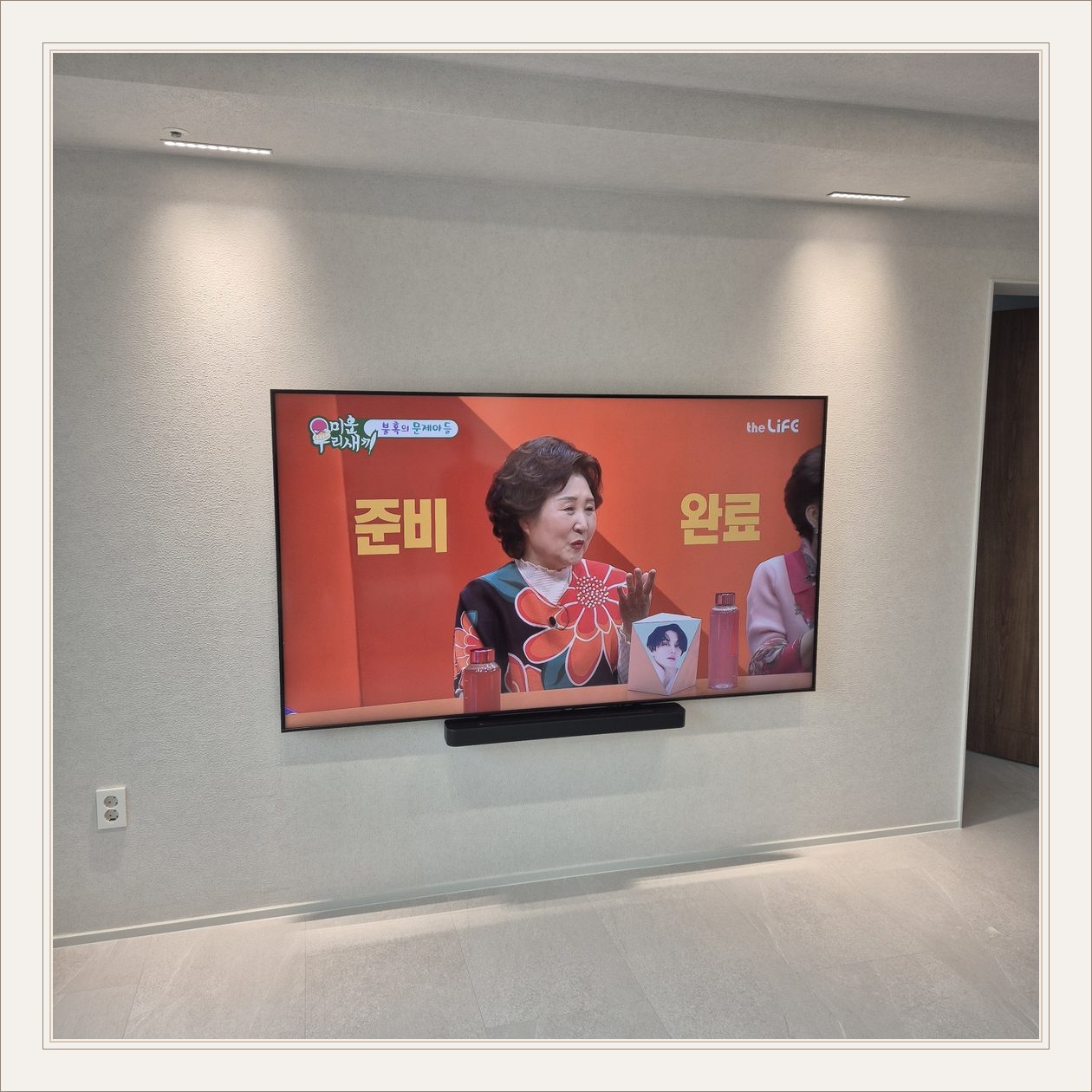 조명이 비추는 미색 벽과 사운드바, 화면 아래 가로로 놓인 검은 음향기기가 선명합니다