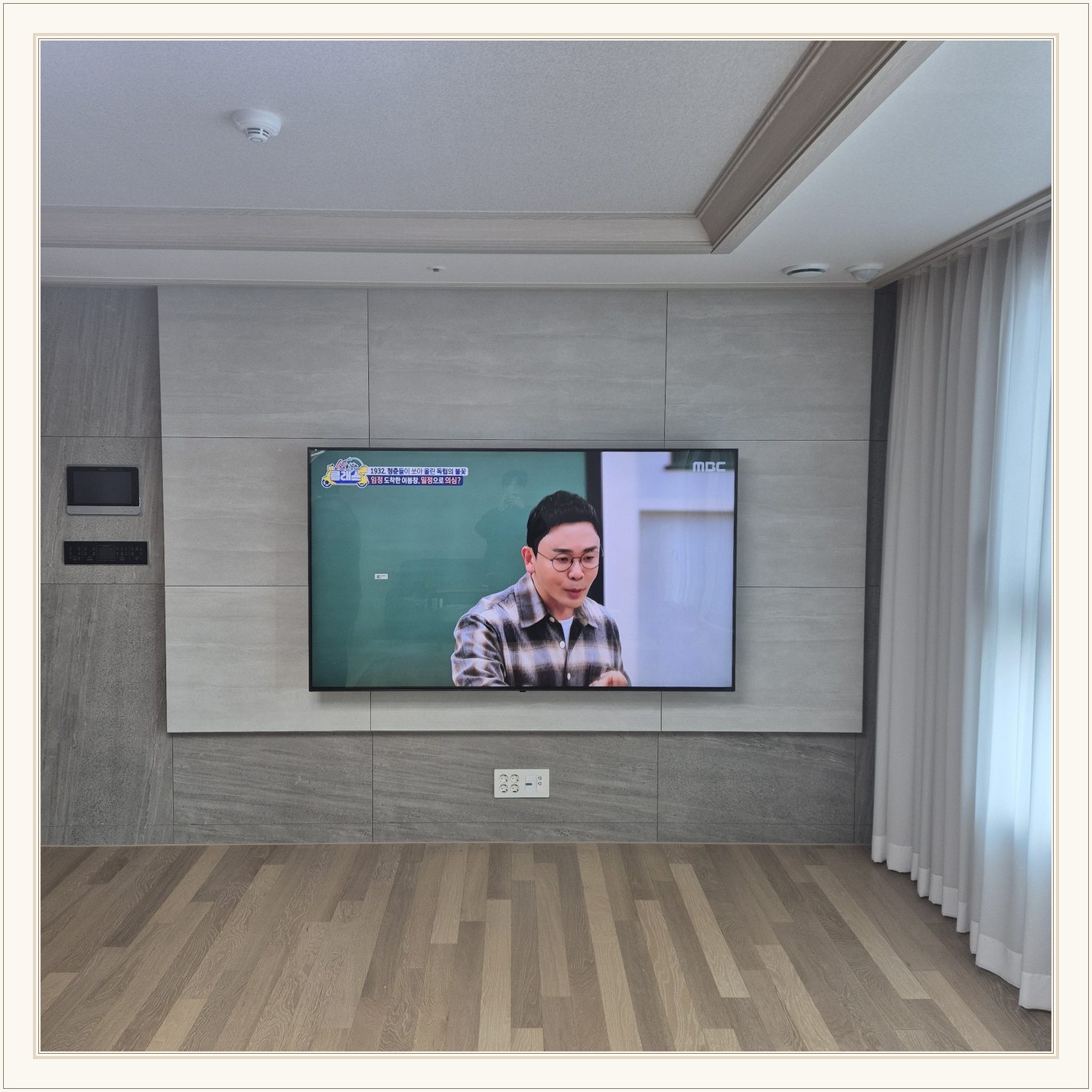거실 회색 패널과 벽걸이 TV, 오른쪽 커튼 옆으로 화면이 중심에 놓여 있습니다