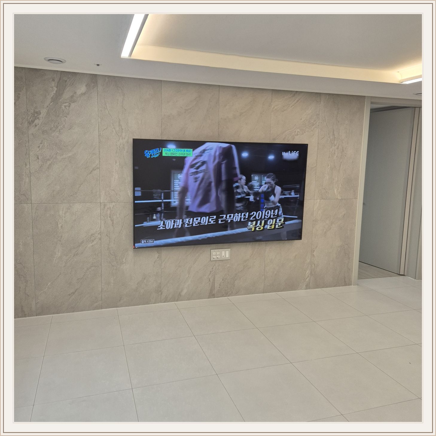 석재 느낌 벽면을 정면에서 본 TV, 넓은 바닥 여유와 조명 라인이 동시에 보입니다