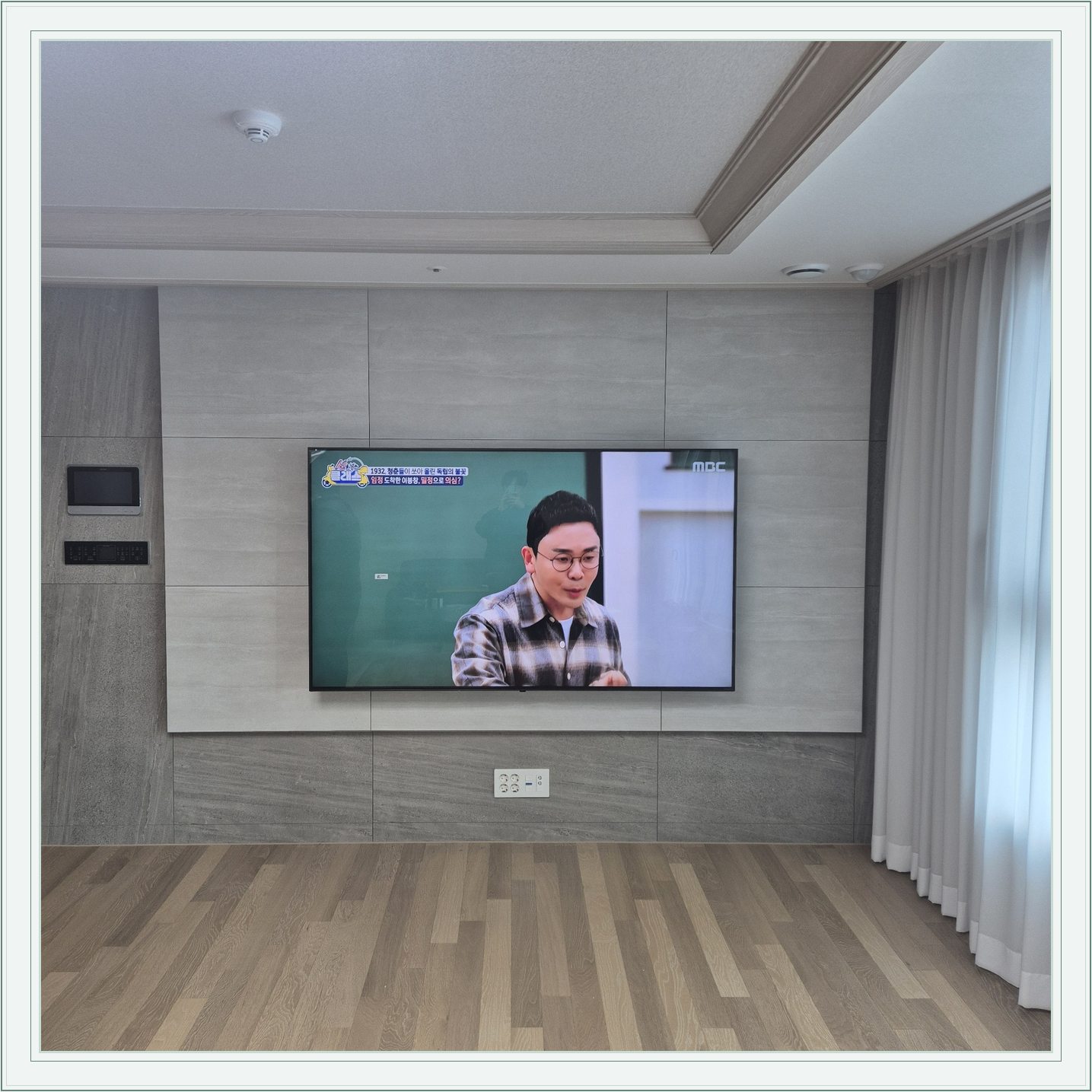 거실 회색 패널과 벽걸이 TV, 오른쪽 커튼 옆으로 화면이 중심에 놓여 있습니다