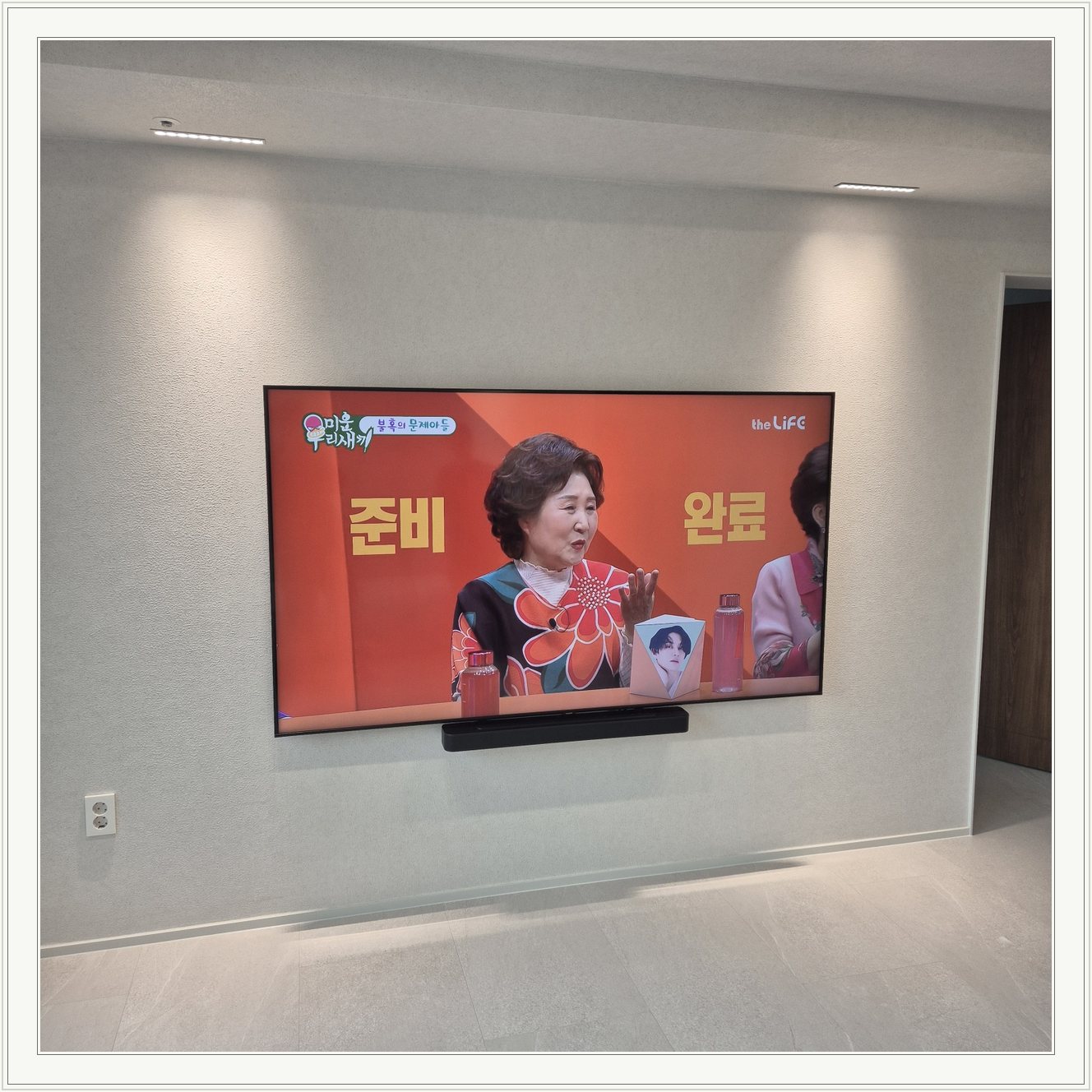 조명이 비추는 미색 벽과 사운드바, 화면 아래 가로로 놓인 검은 음향기기가 선명합니다